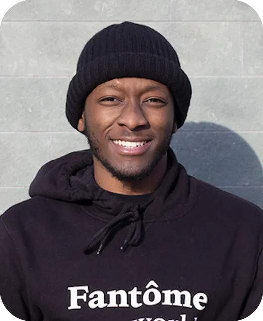 Smiling man wearing a black beanie and black hoodie with the text 'Fantôme' on it, standing against a gray tiled wall.