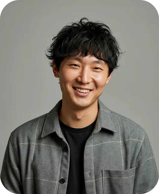 Smiling young man with curly black hair wearing a gray button-up shirt over a black t-shirt against a plain light gray background.