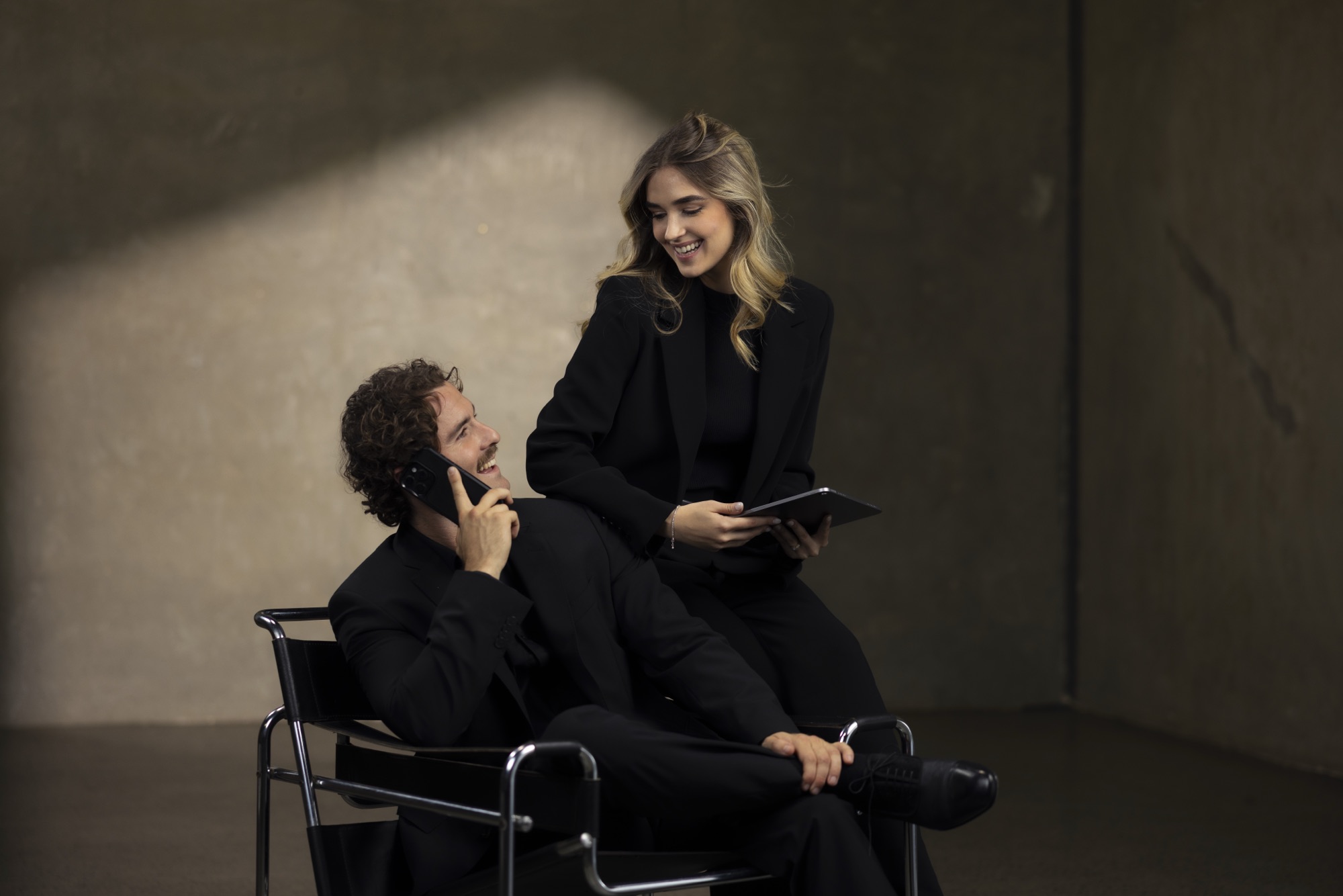 Man on a phone call in a chair while a smiling colleague holds a tablet beside him