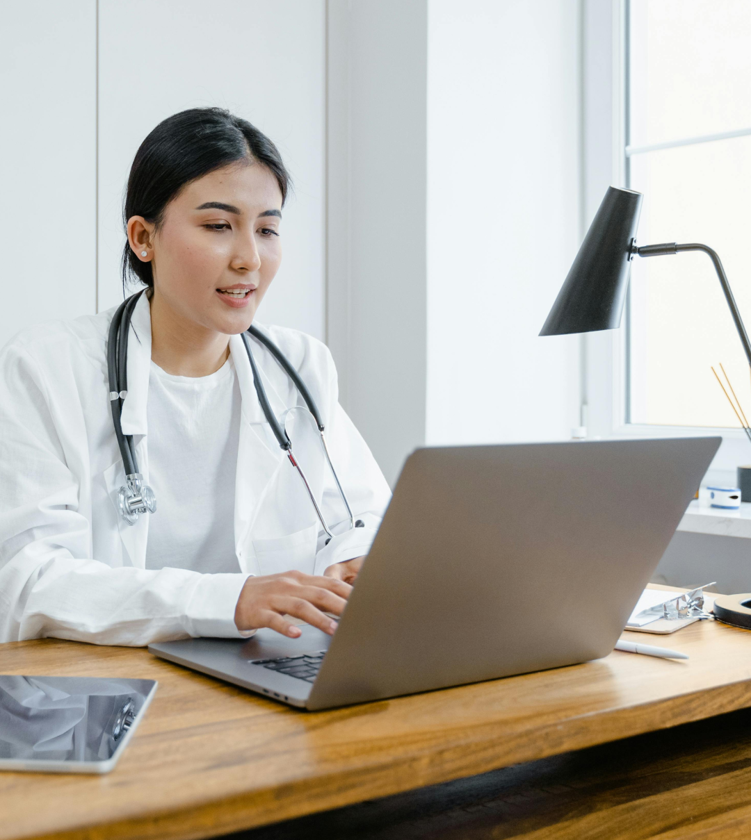 Female doctor wearing white coat and stethoscope typing on a laptop at a wooden desk with a tablet and desk lamp nearby.