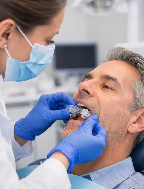 Dentist wearing a mask and blue gloves fits a dental appliance into a male patient's mouth.