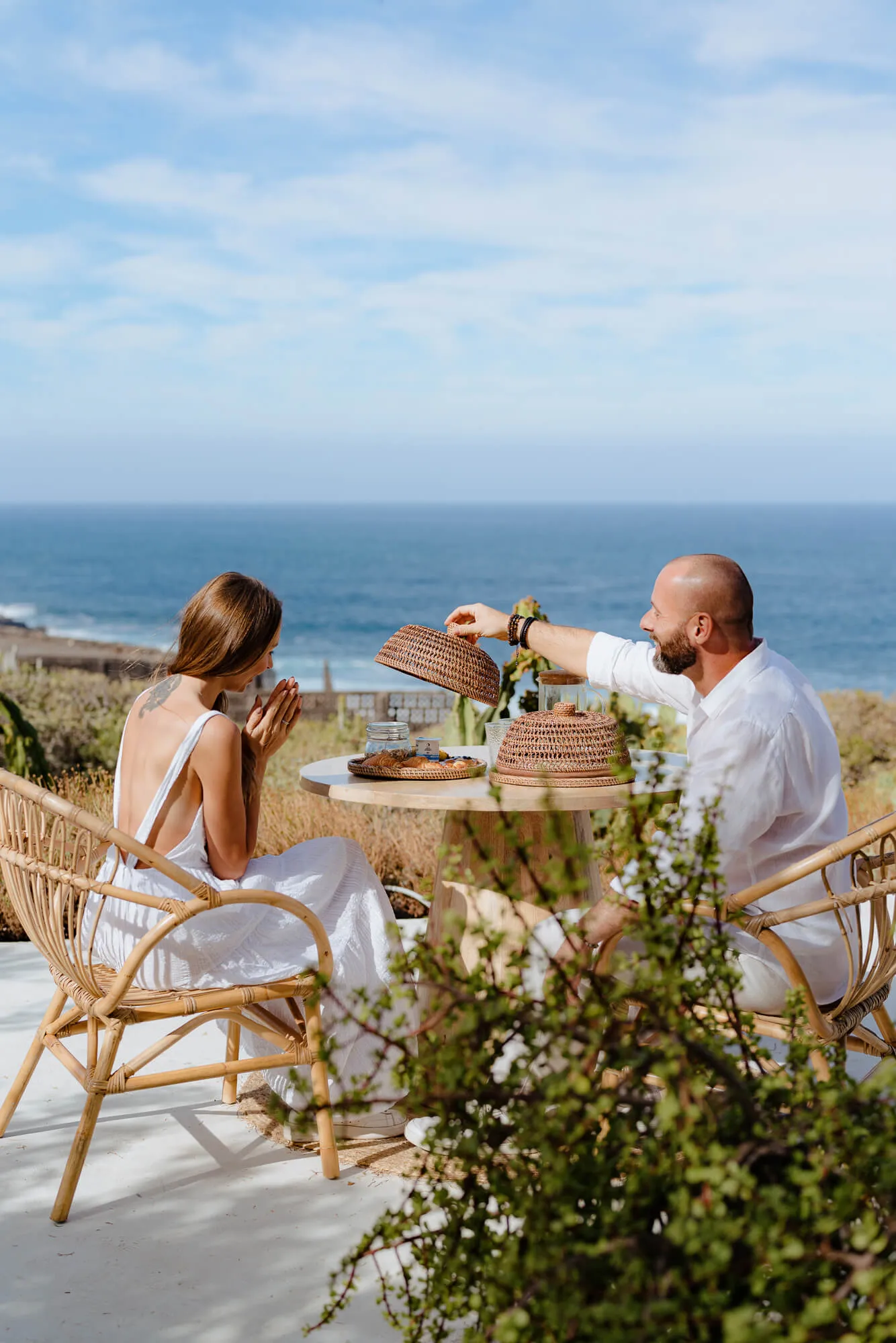 A man and woman sitting at a round table outdoors near the sea; the man lifts a woven food cover while the woman, wearing a white dress, looks on with hands clasped. They sit on rattan chairs with a scenic ocean view and clear sky.