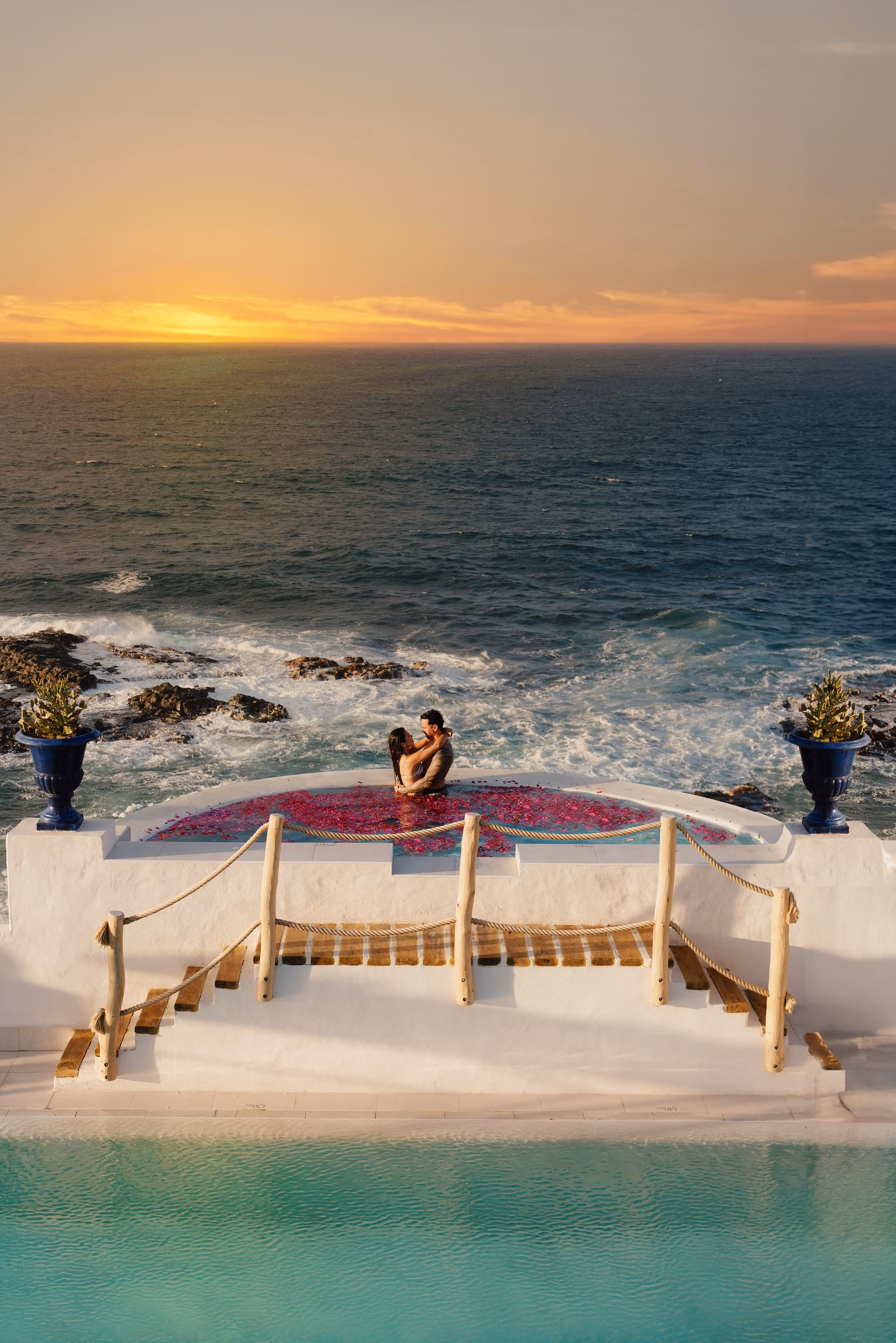 Couple embracing while sitting on a round cushioned platform overlooking the rocky ocean shore at sunset, with a turquoise pool and stairs in the foreground.