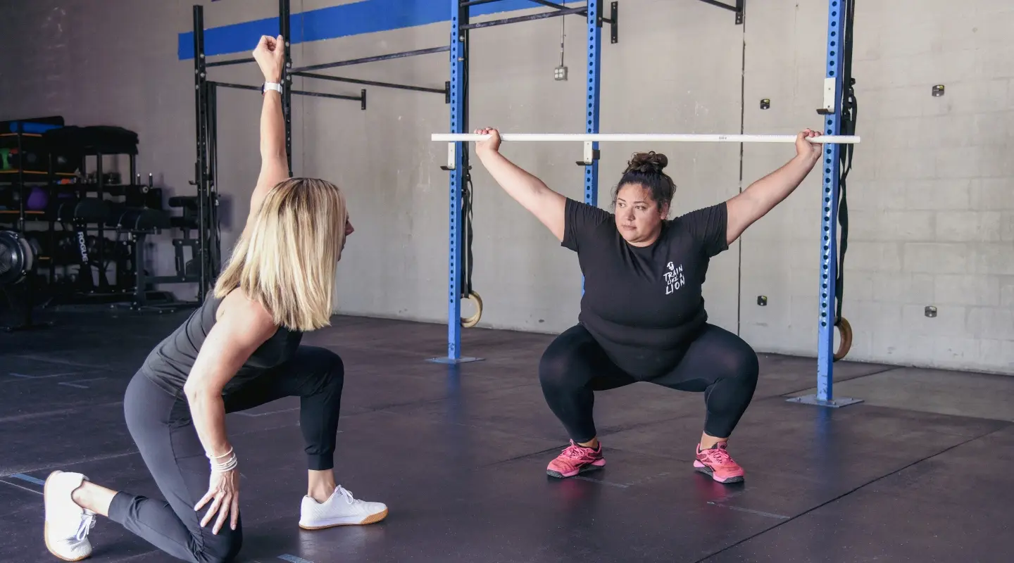 A female personal trainer kneeling to guide a female client through an overhead squat using a PVC pipe at Graham Fitness in Neptune Beach, FL.
