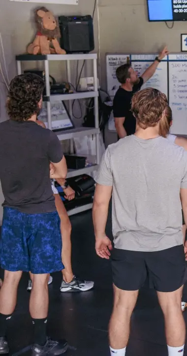 A fitness coach briefing a group of participants on the daily workout using a digital screen and whiteboard during a drop-in class at Graham Fitness in Neptune Beach, FL.