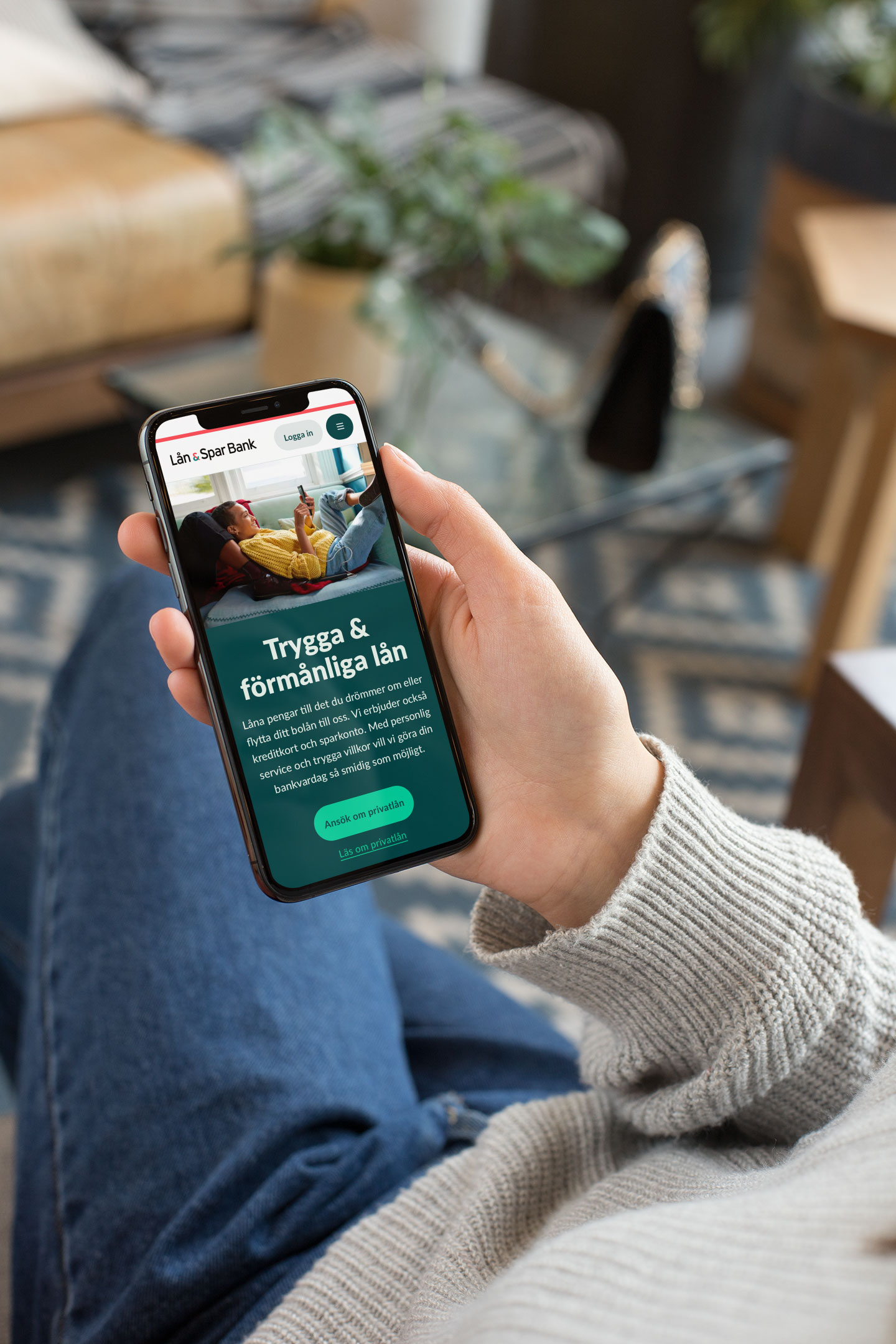 En person i grå tröja och blå jeans håller en smartphone som visar en hemsida för Lån & Spar Bank med text om trygga och förmånliga lån samt knappar för att ansöka om lån.