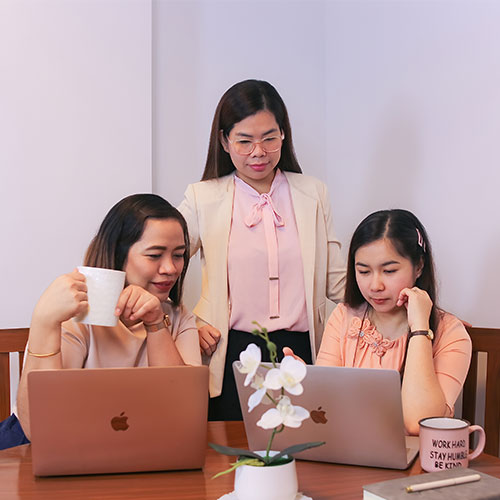 Three women in business attire work together at a table with laptops and coffee mugs.