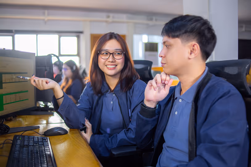 Two colleagues in an office discussing work on a computer screen, with one pointing at the monitor and smiling.