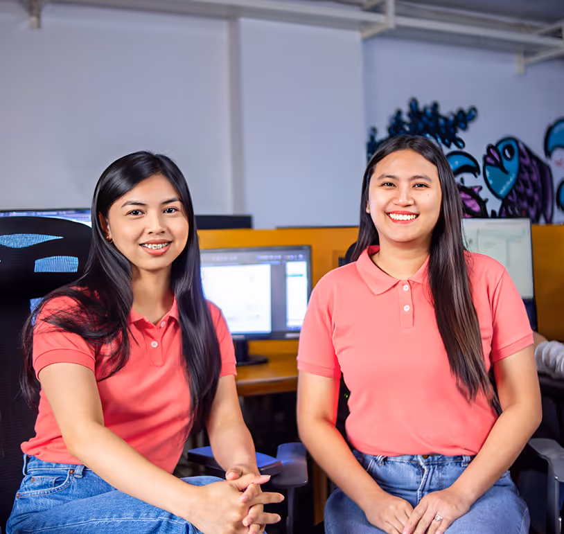 Two young women wearing pink polo shirts sitting and smiling in front of computer monitors in an office.