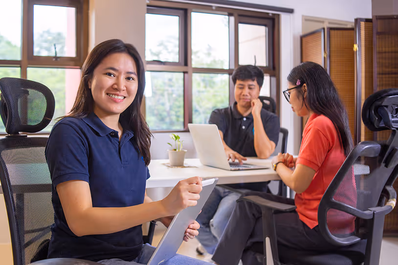 Three people working in an office, with a smiling woman in navy polo holding a tablet in the foreground and two colleagues using a laptop behind her.