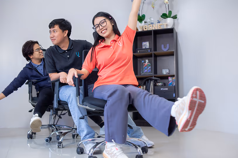 Three young people smiling and leaning back while sitting on office chairs in a playful indoor setting.