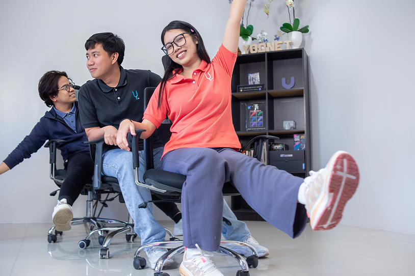 Three young people smiling and leaning back while sitting on office chairs in a playful indoor setting.