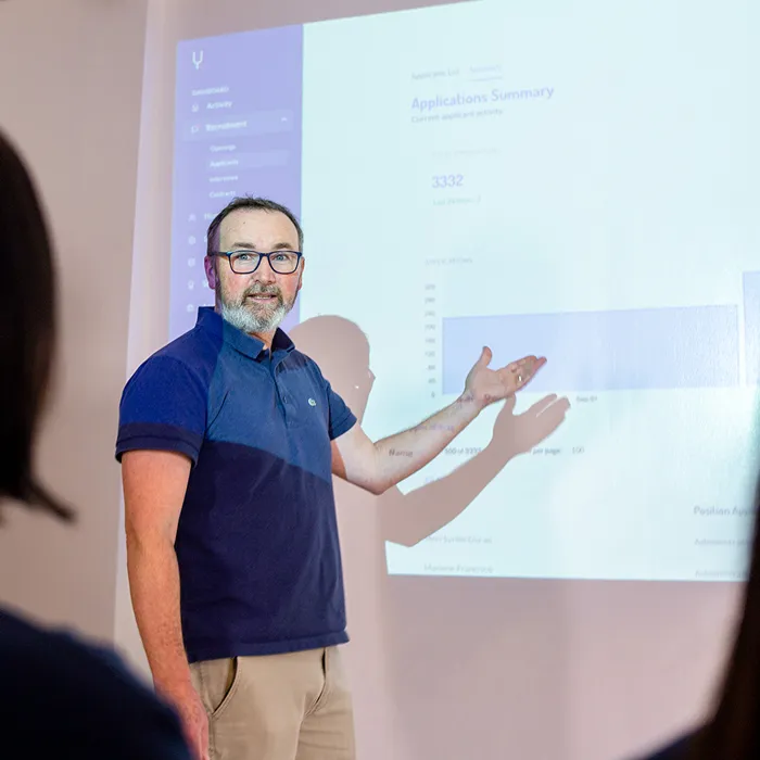 Man with glasses pointing at a projected application summary chart during a presentation to an audience.
