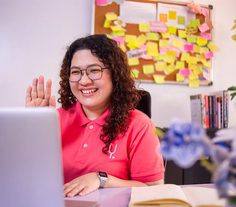 Smiling woman with curly hair and glasses waving during a video call on a laptop in a cozy office.