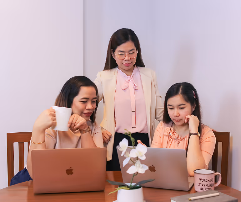 Three women working with two laptops at a wooden table, one holding a white mug and another with a pink mug that reads 'WORK HARD STAY HUMBLE BE KIND'.