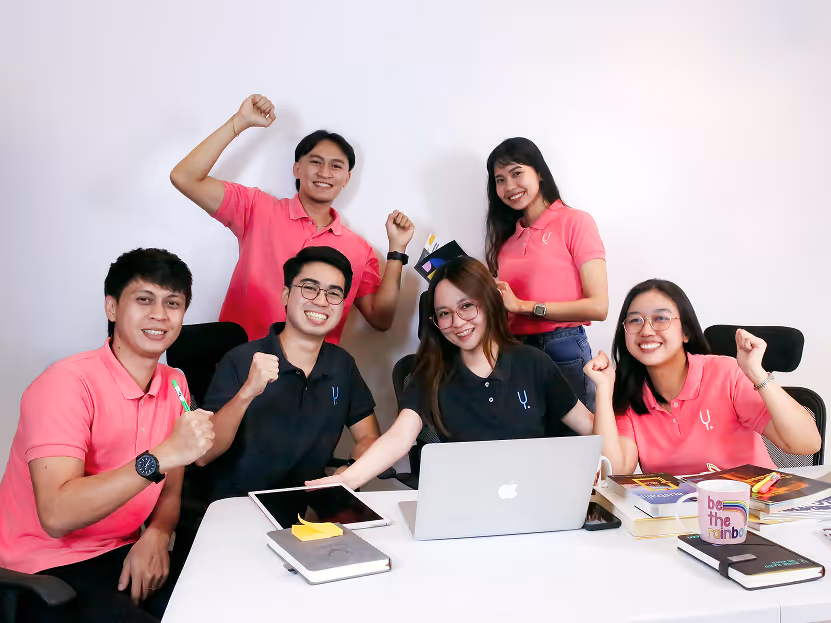 Six smiling colleagues cheering around a laptop and tablet at a white office table.