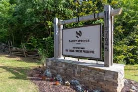 Sandy Springs Big Trees Forest Preserve entrance sign mounted on wooden posts with greenery and a wooden fence in the background.