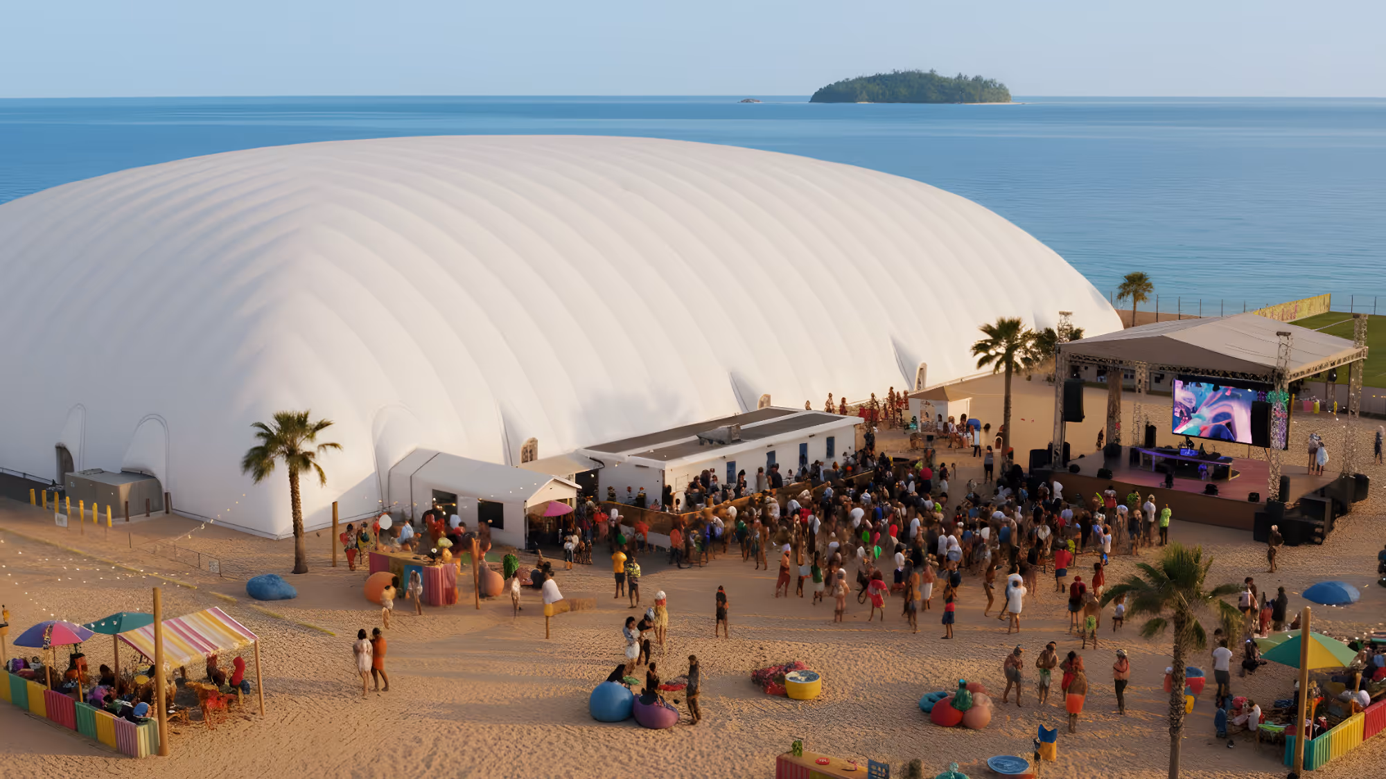 Outdoor event on sandy beach with a large white air dome, crowd of people, stage with screen, palm trees, and a calm ocean with a small island in the background.