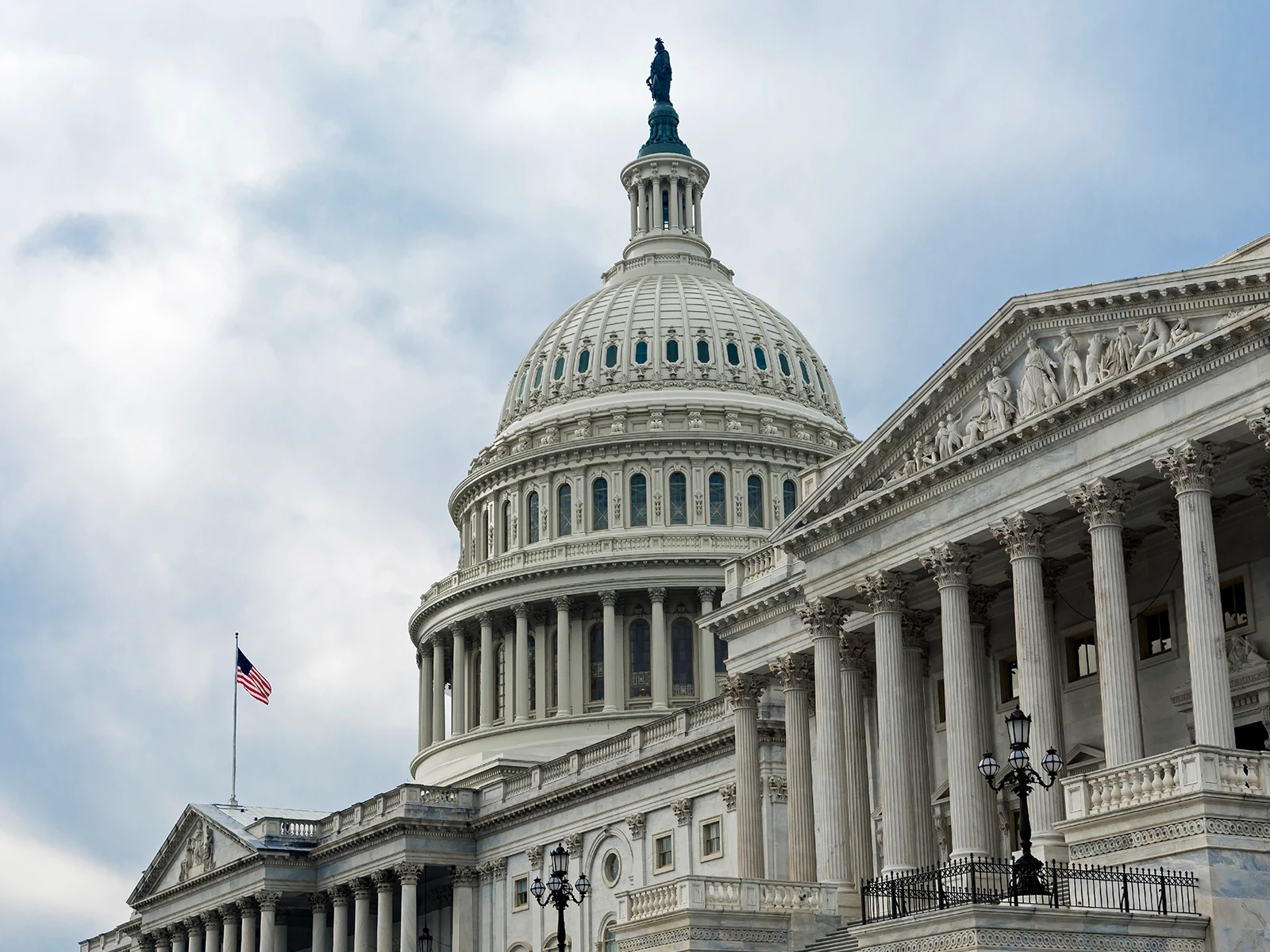 The U.S. Capitol building with its dome, detailed columns, and an American flag against a cloudy sky.