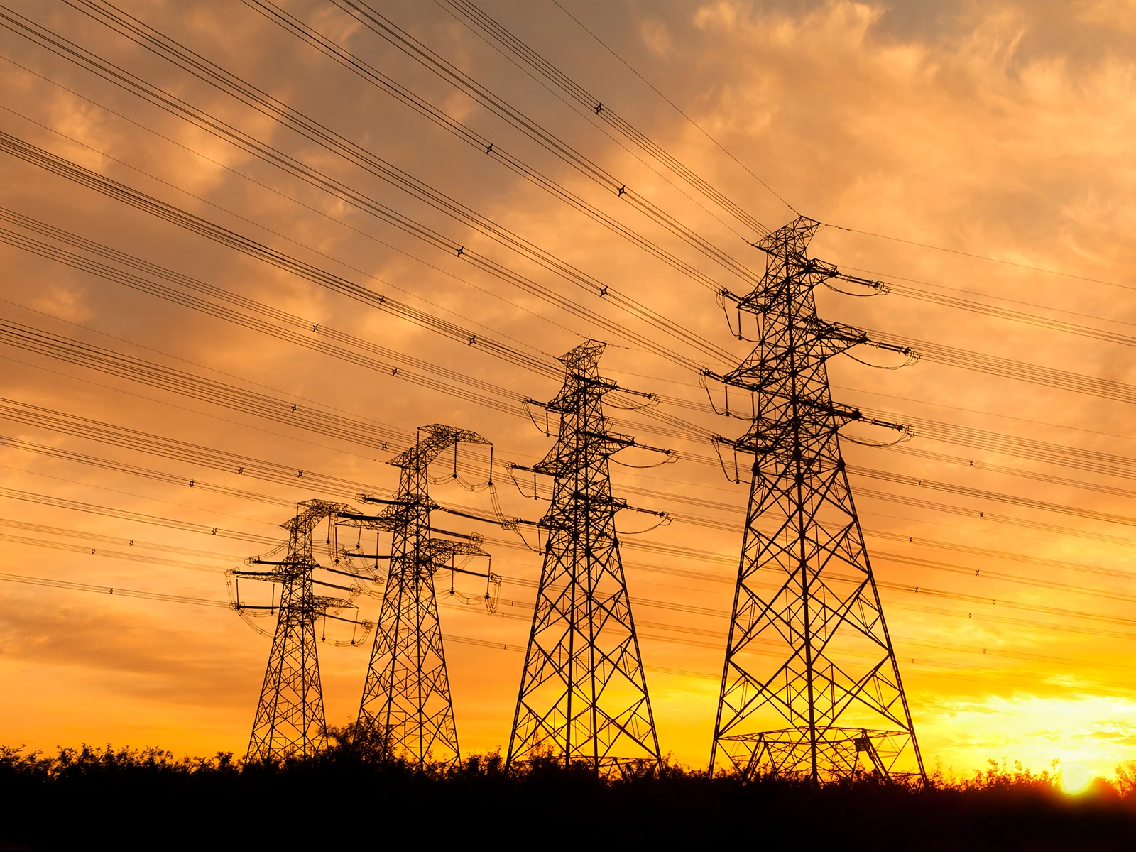 夕暮れ時、木々のシルエットを背景に、送電線が張られた4本の大きな送電塔が浮かび上がっている。
