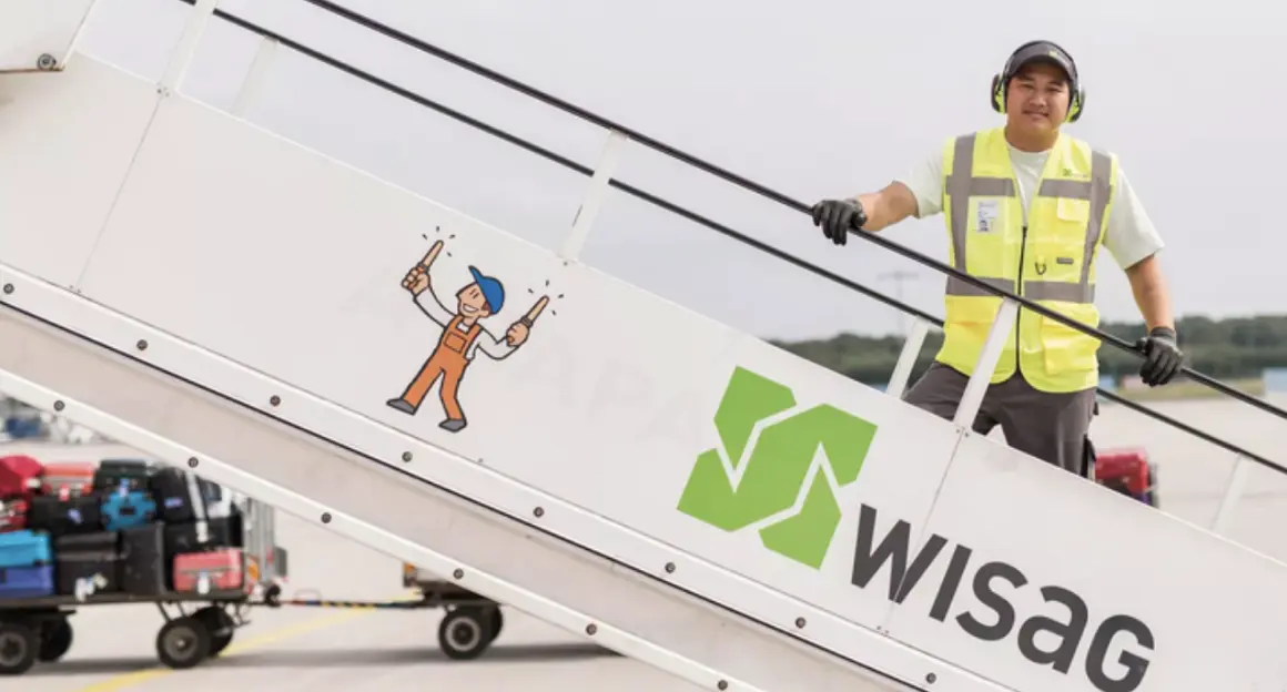 An airport ground staff wearing a yellow high-visibility vest, standing on the steps of mobile aircraft stairs. The side of the stairs features the green WISAG logo.