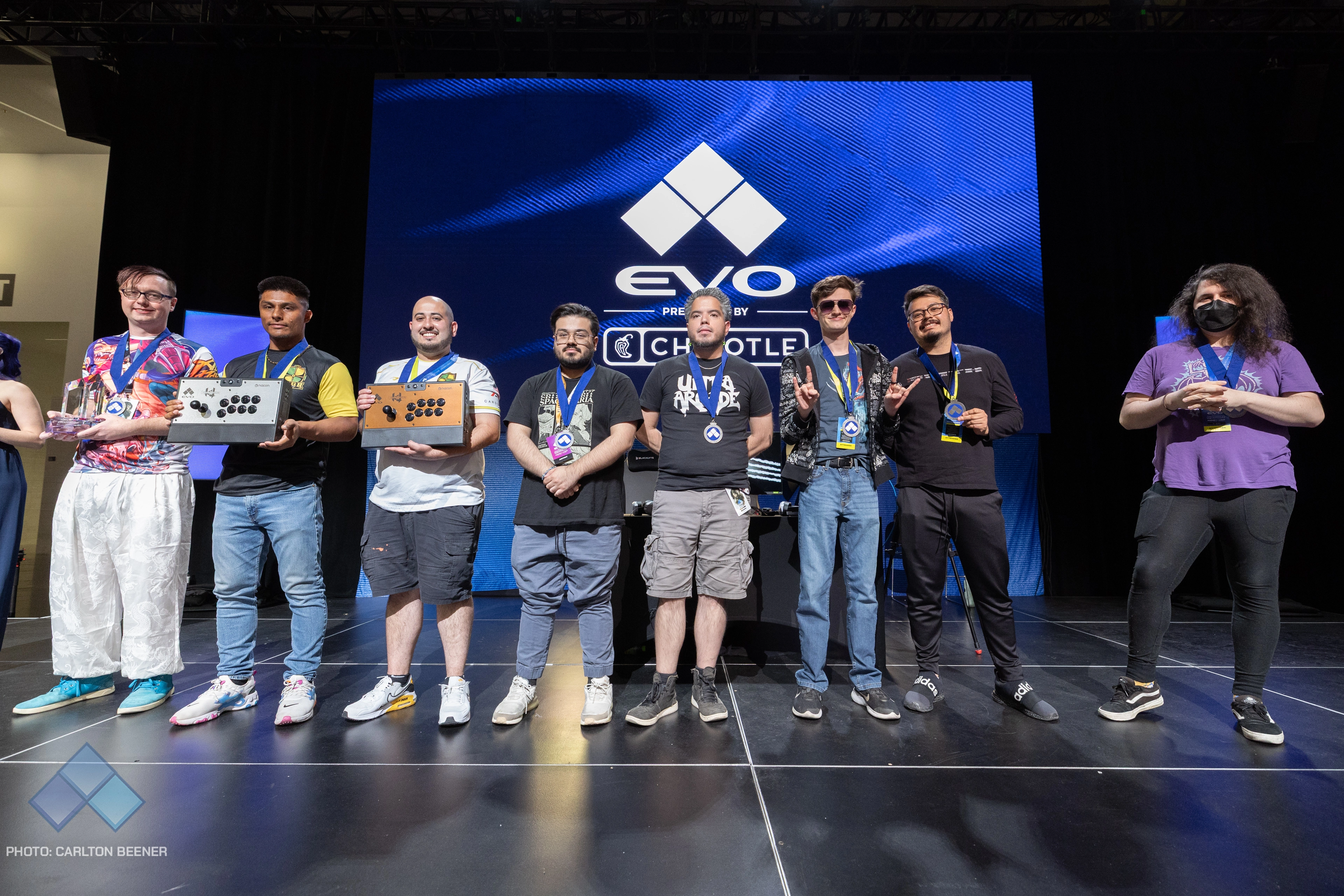 Evo's KI Top 8 on stage, posing with their medals, fight sticks, and trophies