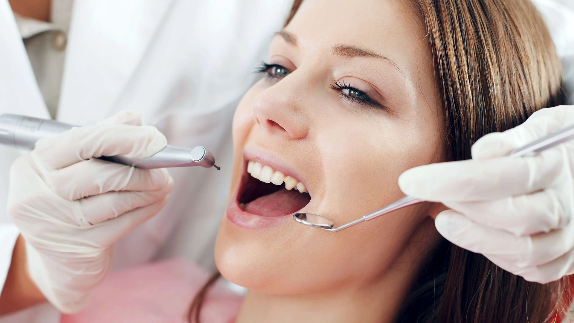Dentist wearing white gloves examining a smiling woman's open mouth using dental tools.