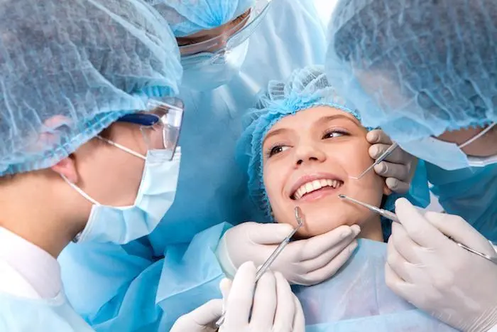 Smiling patient undergoing dental examination by three dentists wearing masks, gloves, and surgical caps.