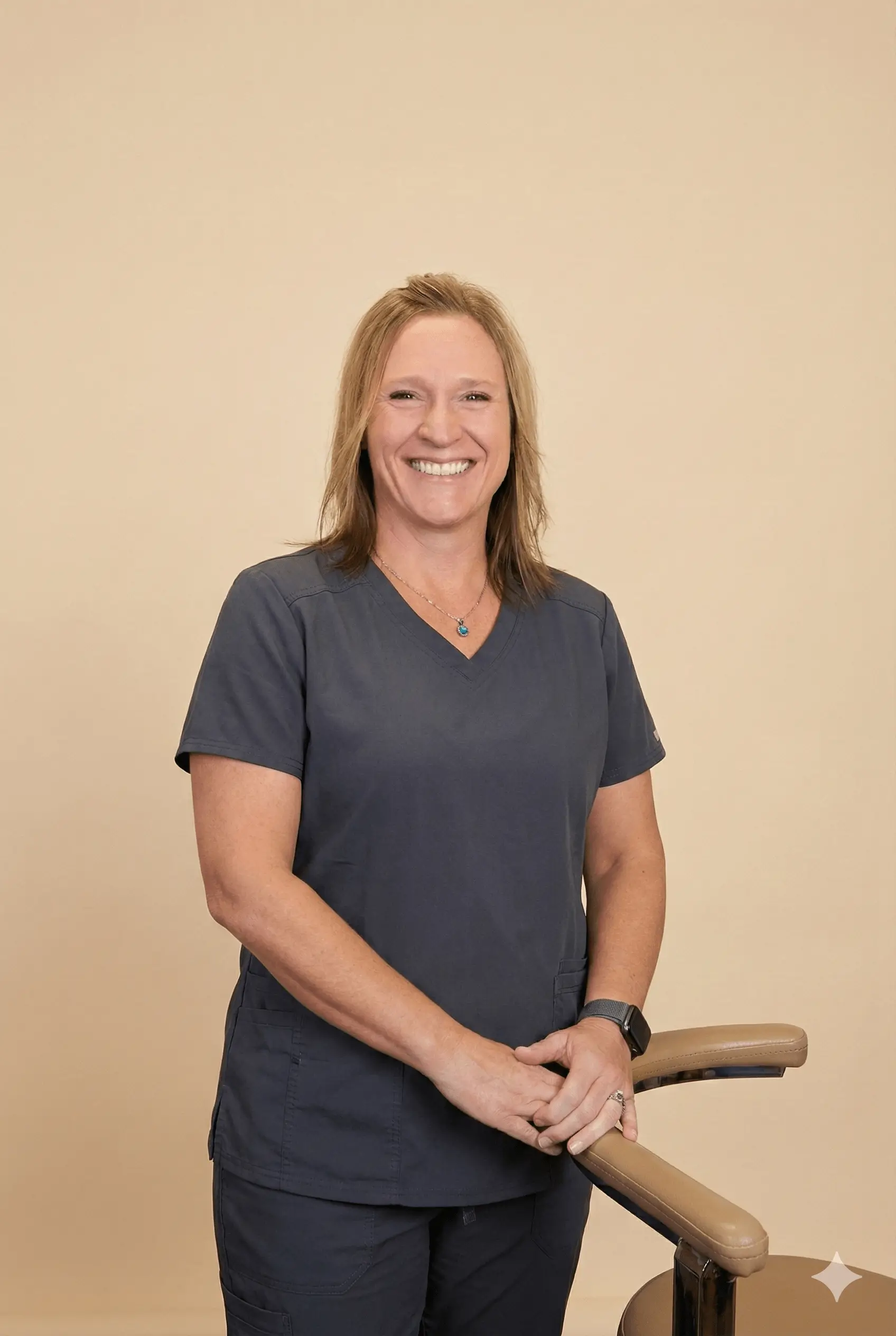 Smiling woman in dark blue scrubs standing with hands resting on a beige padded chair arm against a light tan background.