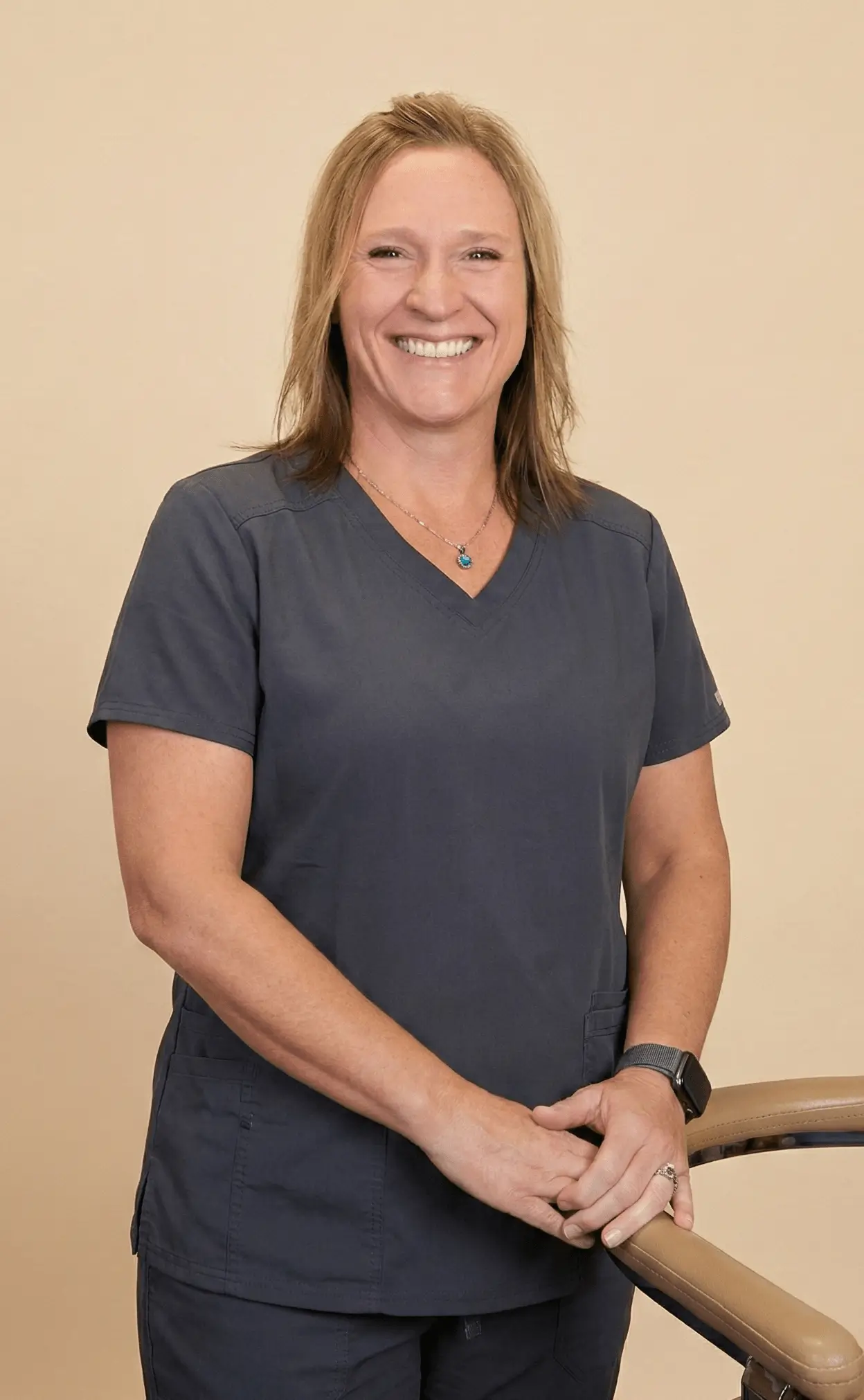 Smiling woman in navy blue medical scrubs standing with hands resting on a chair armrest against a beige background.