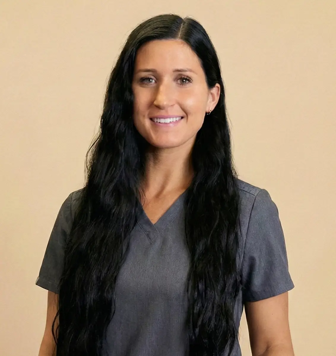 Smiling woman with long black hair wearing a gray V-neck shirt against a beige background.