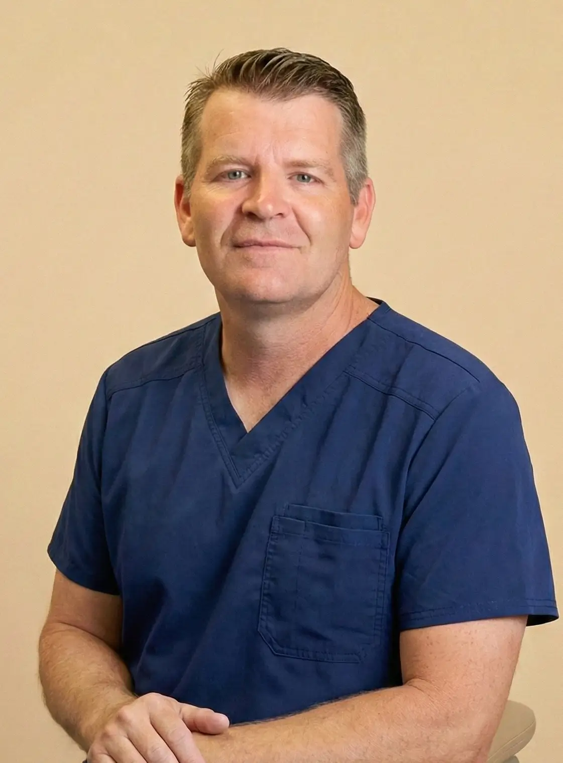 Dr. Jerry Walker, DMD with short brown hair wearing a navy blue scrub top sitting with hands clasped.