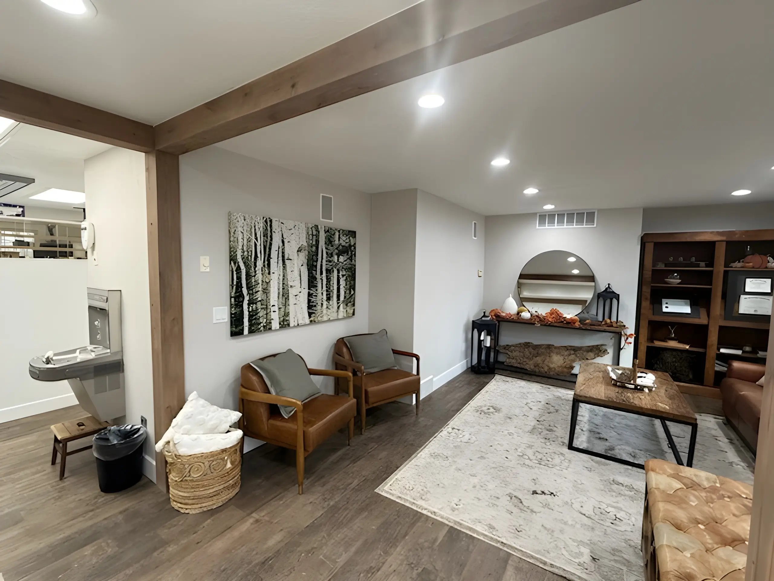 Cozy modern office waiting area with two leather chairs, a basket with blankets, a wooden table, a round mirror, and decorative items.