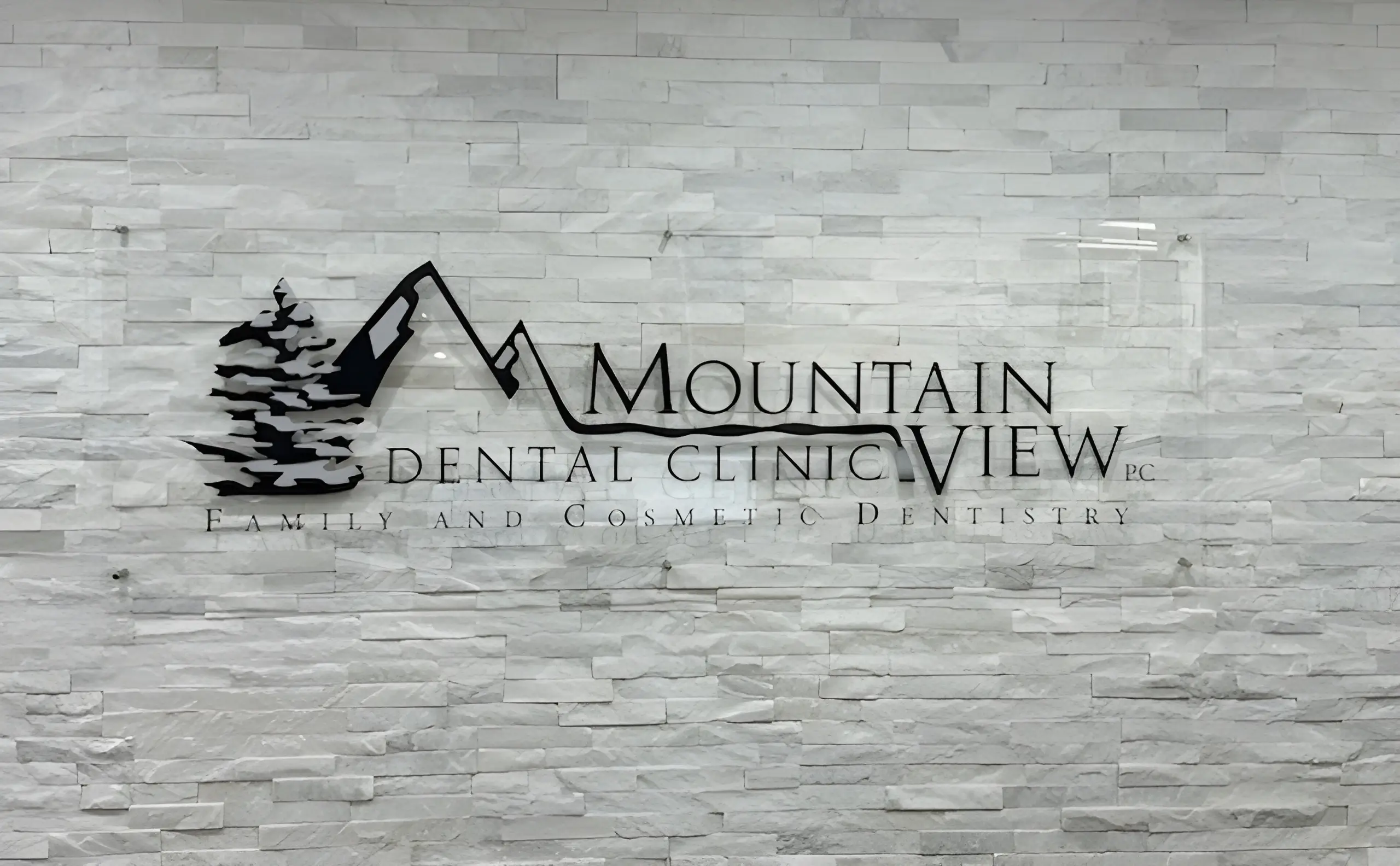 Glass sign for Mountain View Dental Clinic with tree and mountain graphic on white stone wall.
