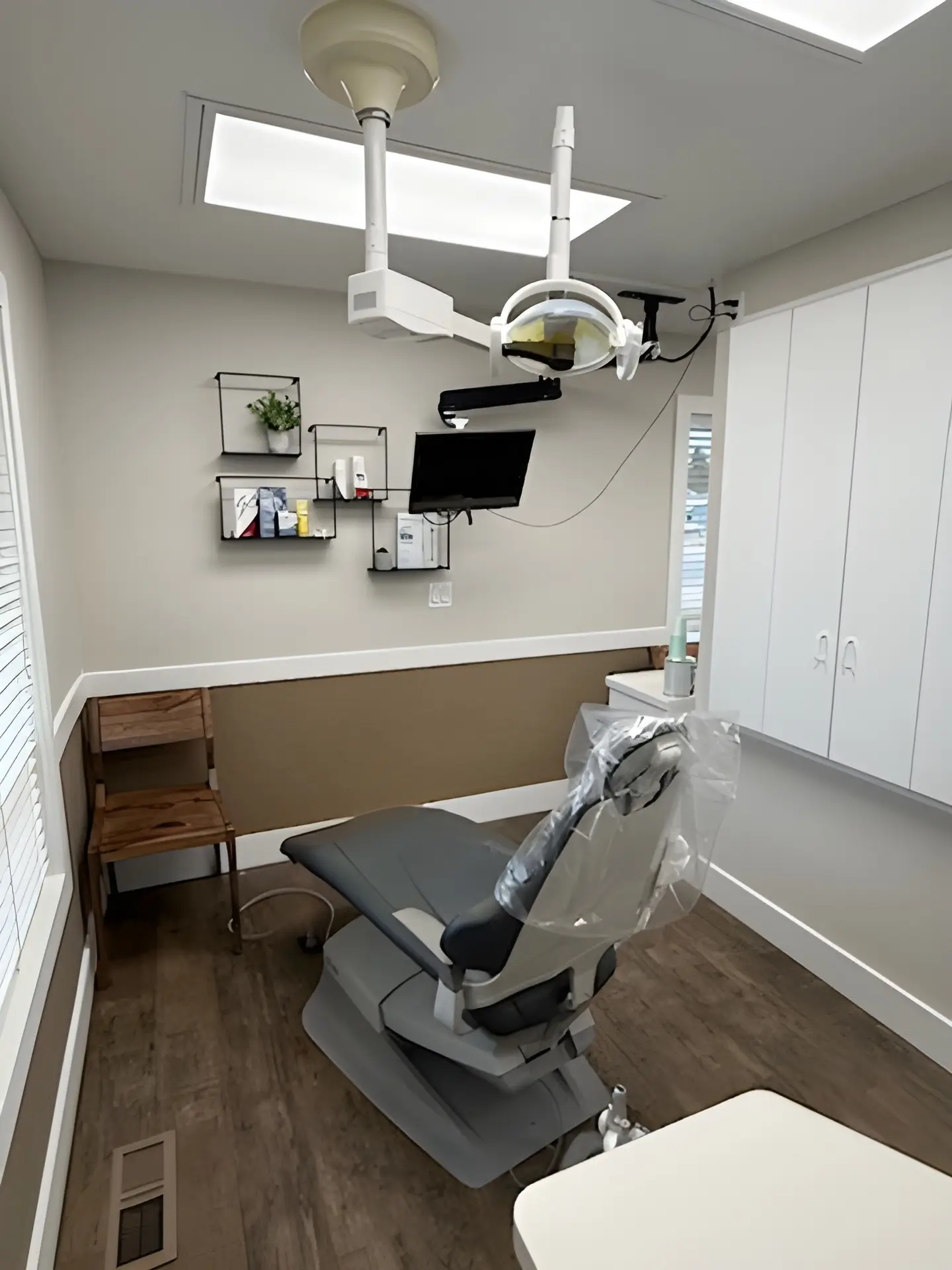 Modern dental examination room with dental chair, overhead light, wall-mounted monitor, wooden chair, and shelves with dental supplies.