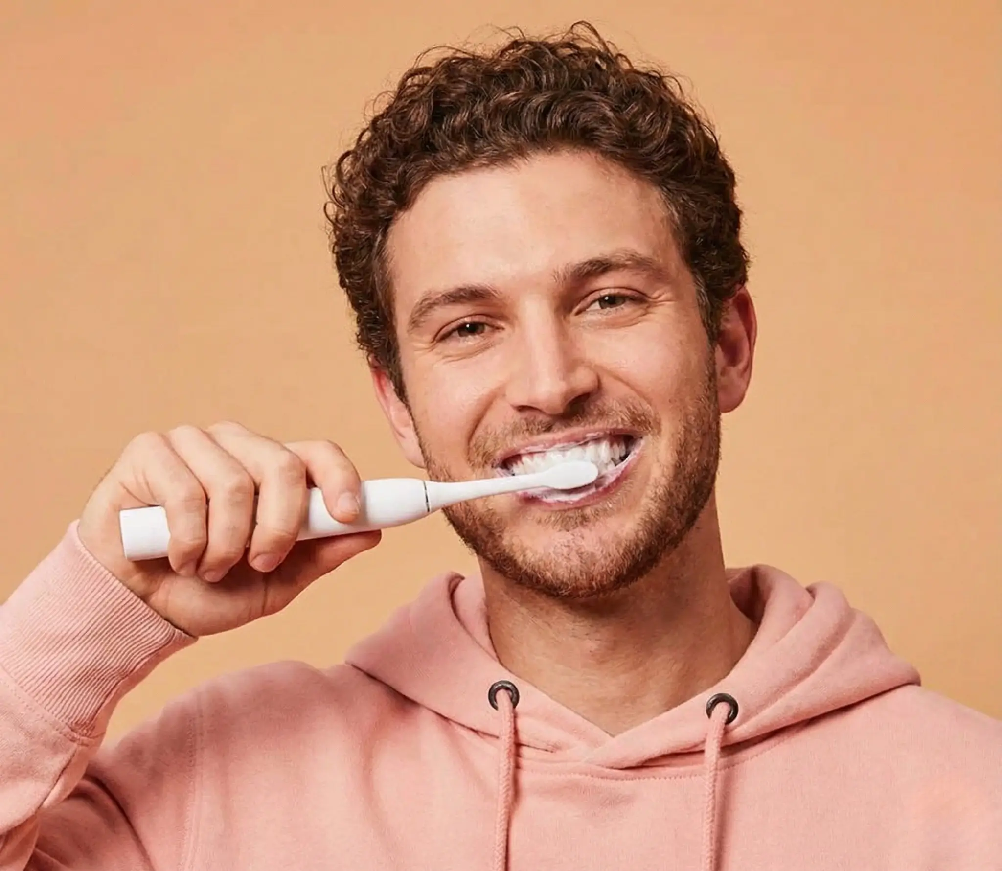 Man with curly hair smiling while brushing his teeth with an electric toothbrush and wearing a pink hoodie.