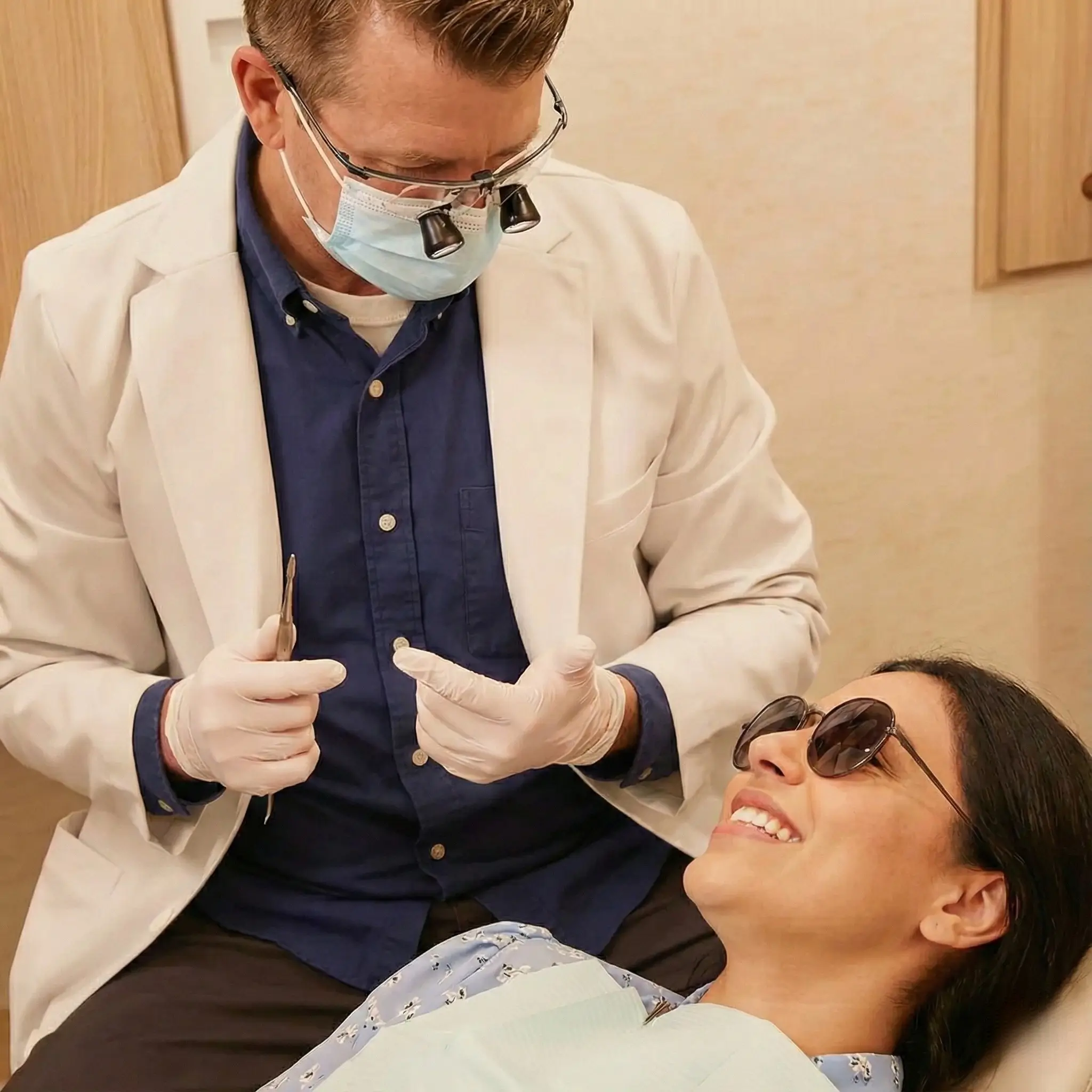Dentist wearing magnifying loupes and gloves examines smiling female patient reclining in dental chair with sunglasses.