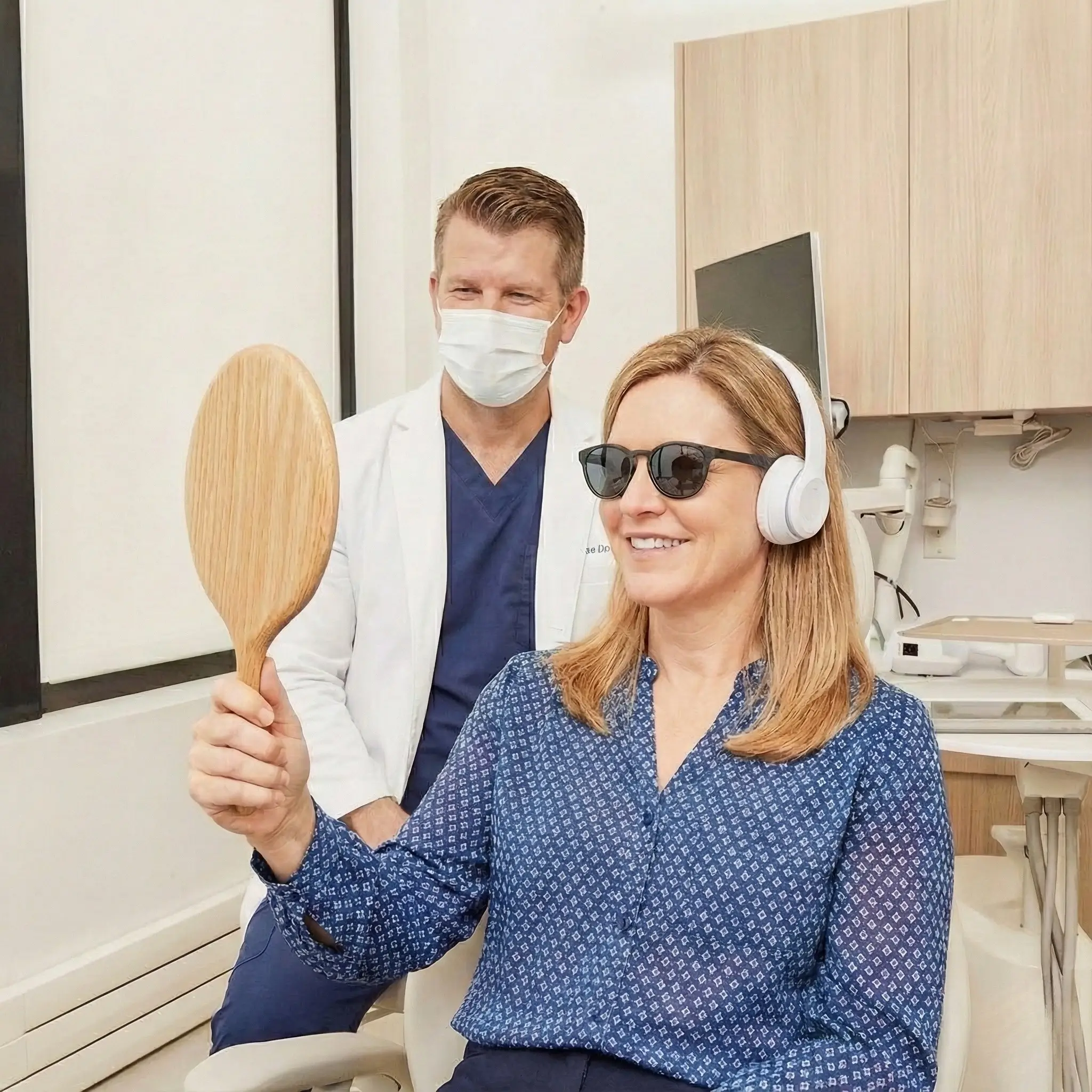 Woman wearing sunglasses and headphones smiles while holding a wooden hand mirror in a medical office, with a masked male doctor in a white coat behind her.