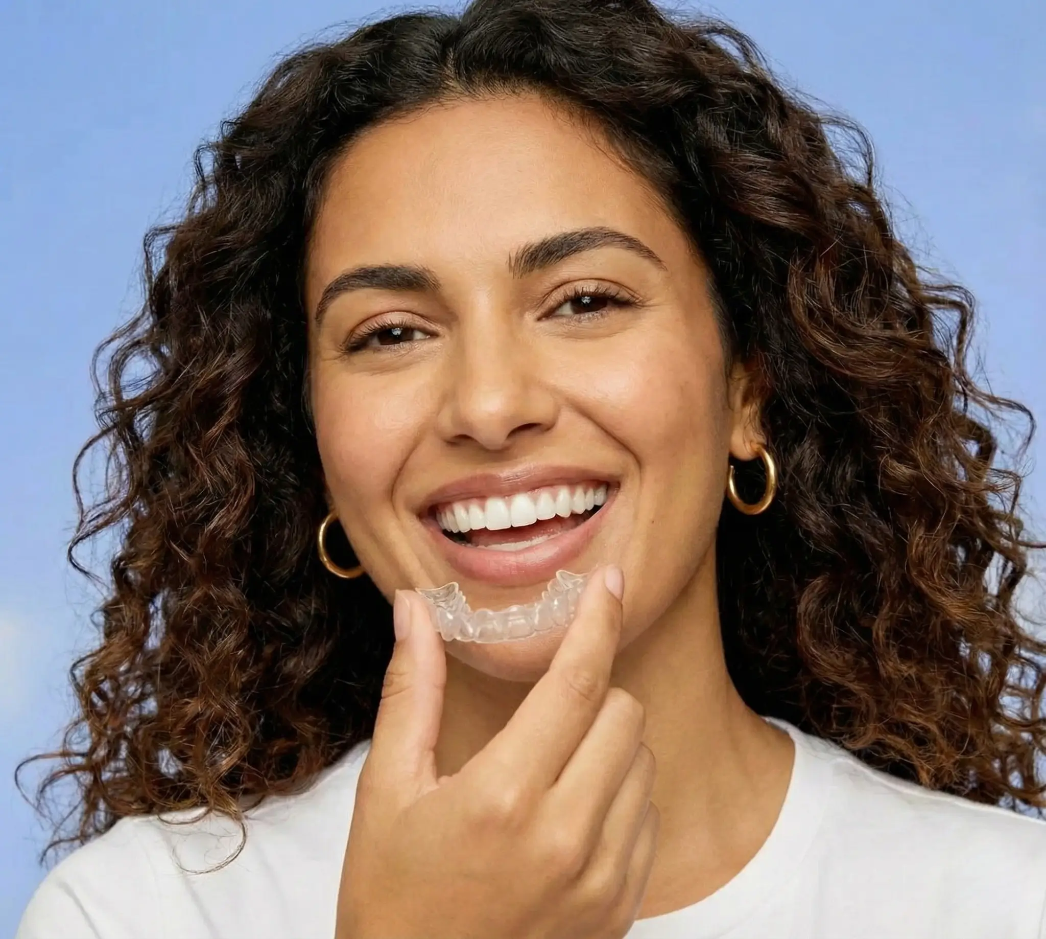 Smiling woman with curly hair holding a clear dental aligner near her mouth.