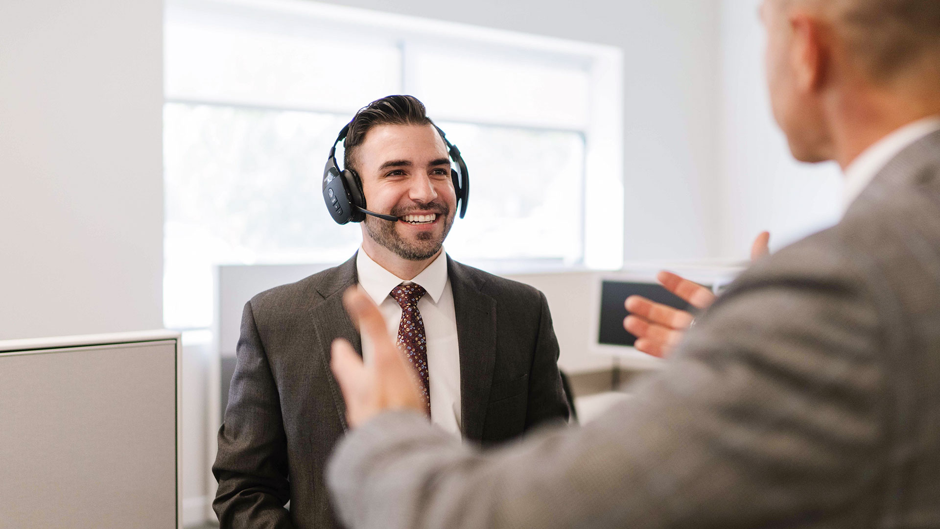 A man smiles while talking to another person, he is wearing a headset.