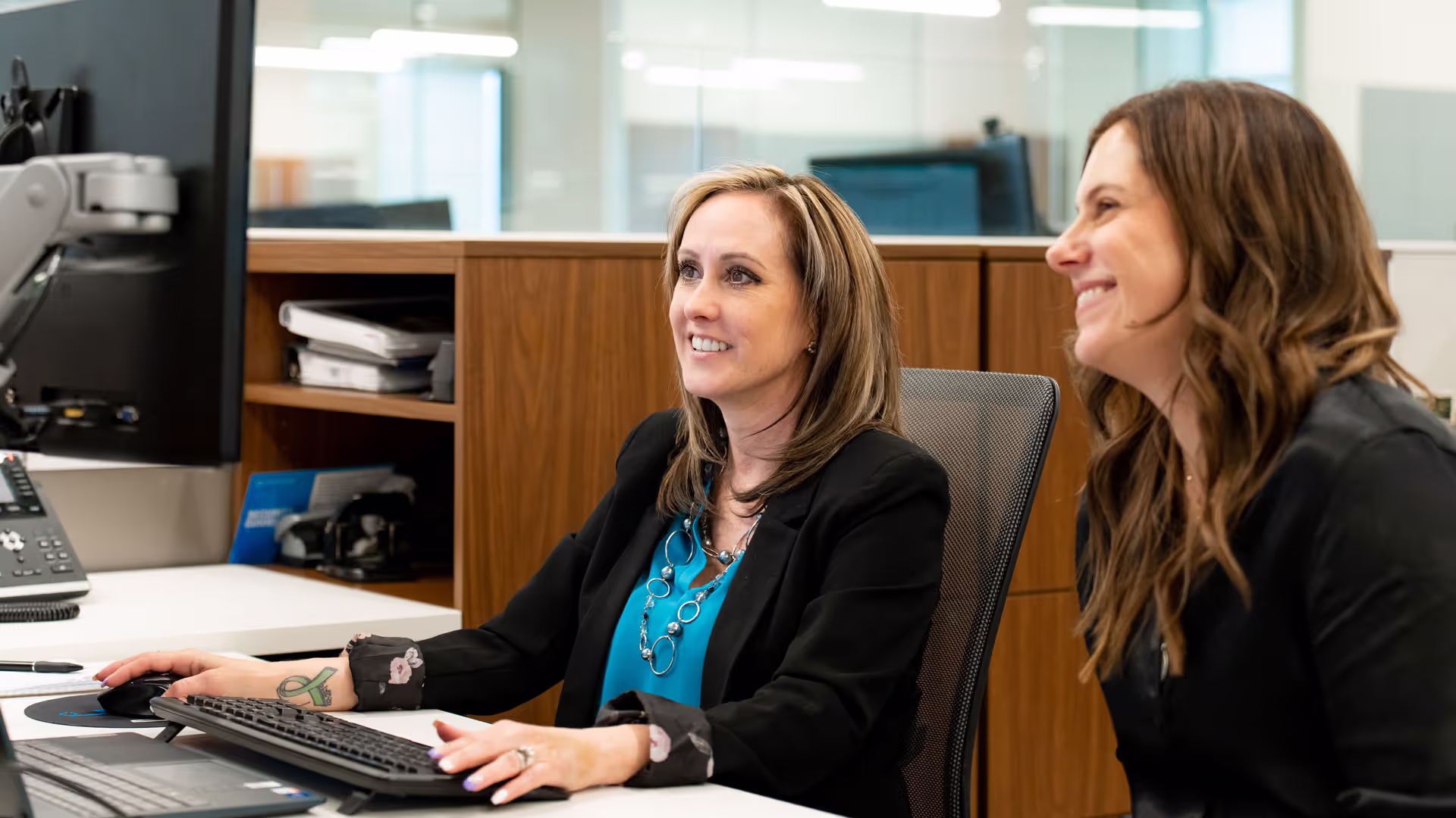 Two businesswoman sitting at a desk together smiling and looking at a computer screen in a modern office space
