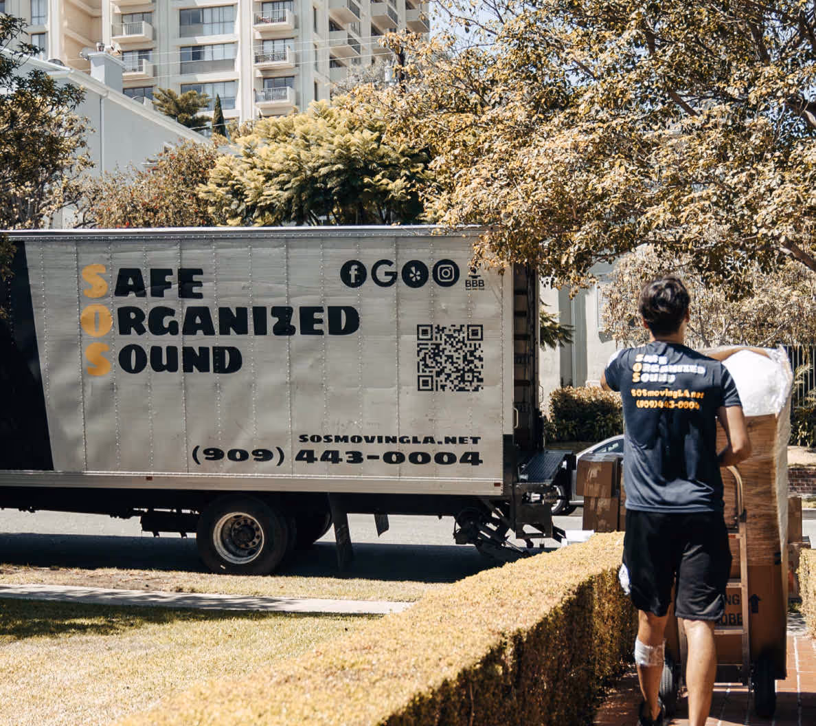 Person pushing a dolly with boxes near a moving truck labeled SOS Moving LA with contact information.