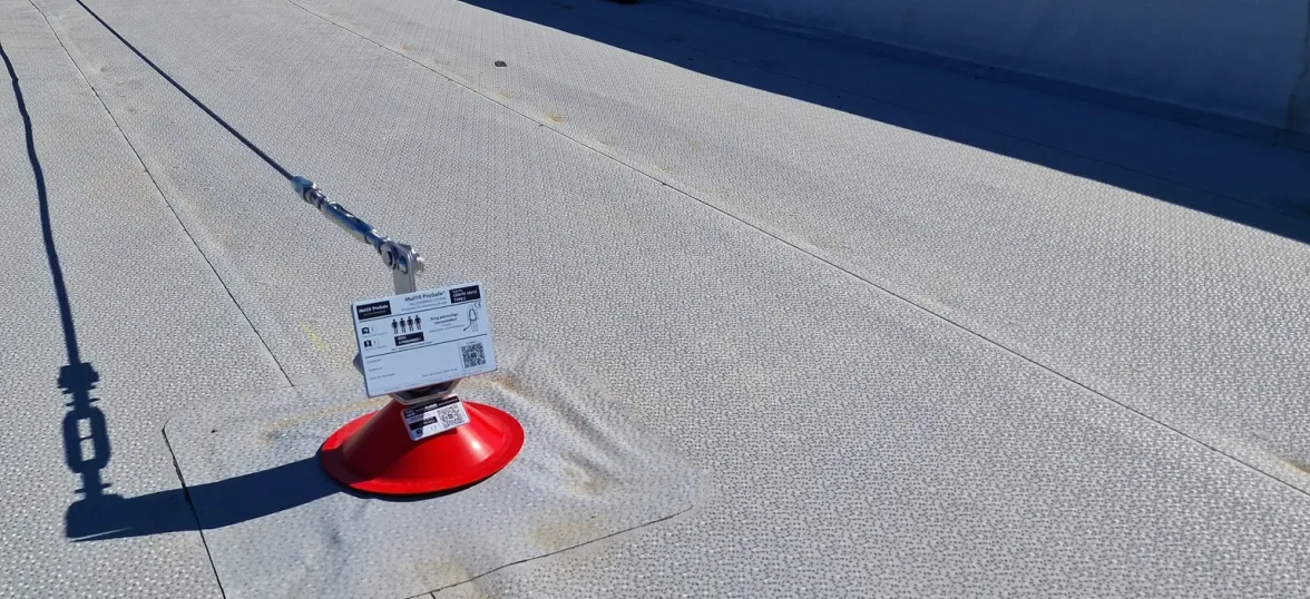 Close-up of a rooftop anchor point with a red base, secured cable, and attached safety instruction tag on a textured roof surface.