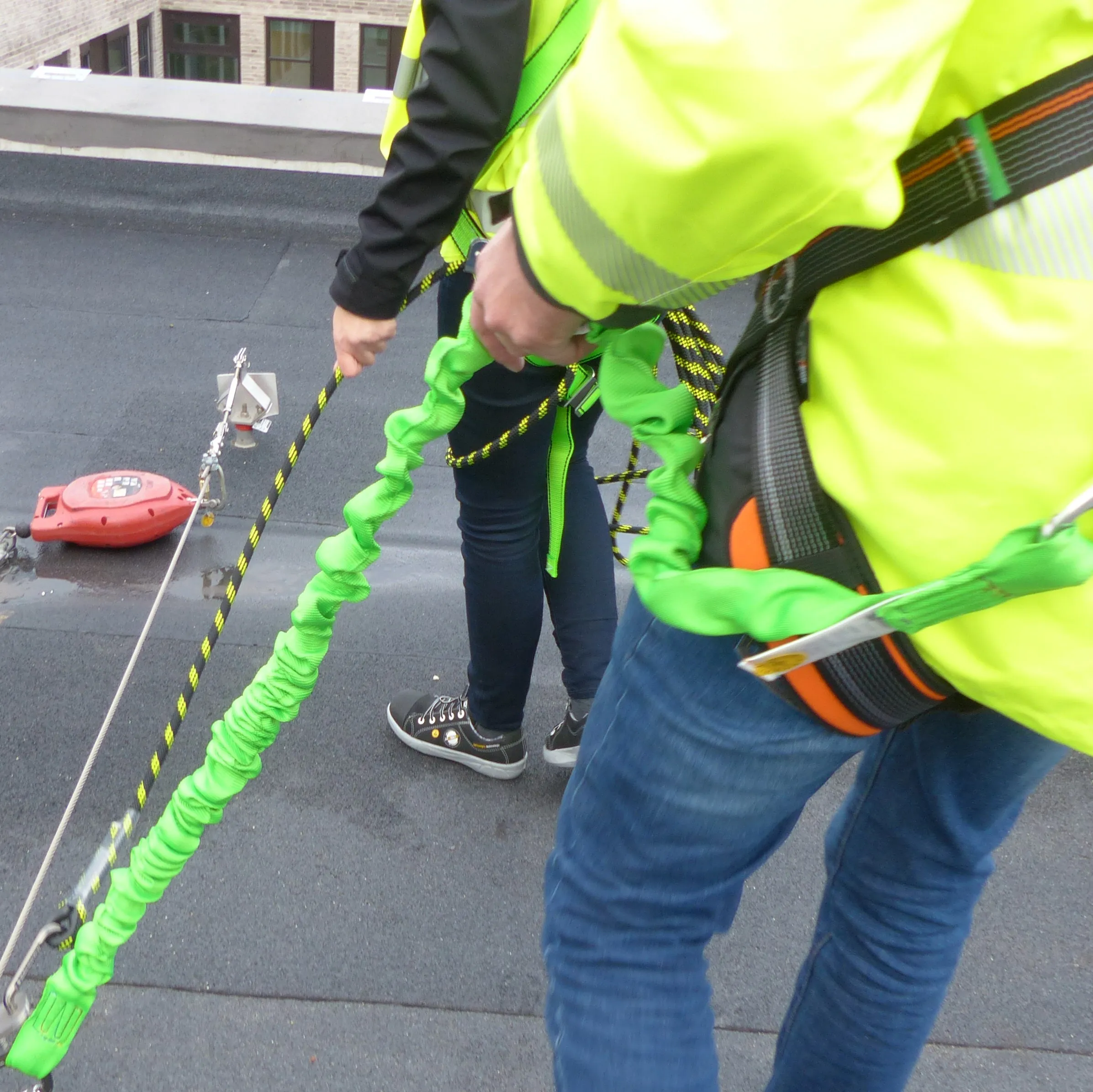 Two workers wearing safety harnesses handling green and black safety ropes on a rooftop.