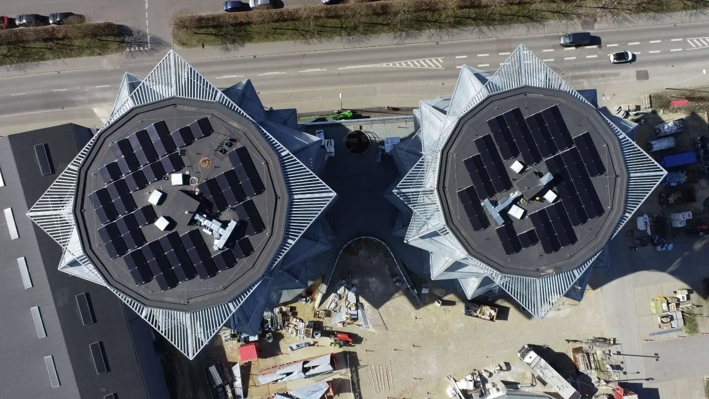Luftfoto af moderne byggeri med to ottekantede tage med solceller og byggeplads mellem bygningerne.
