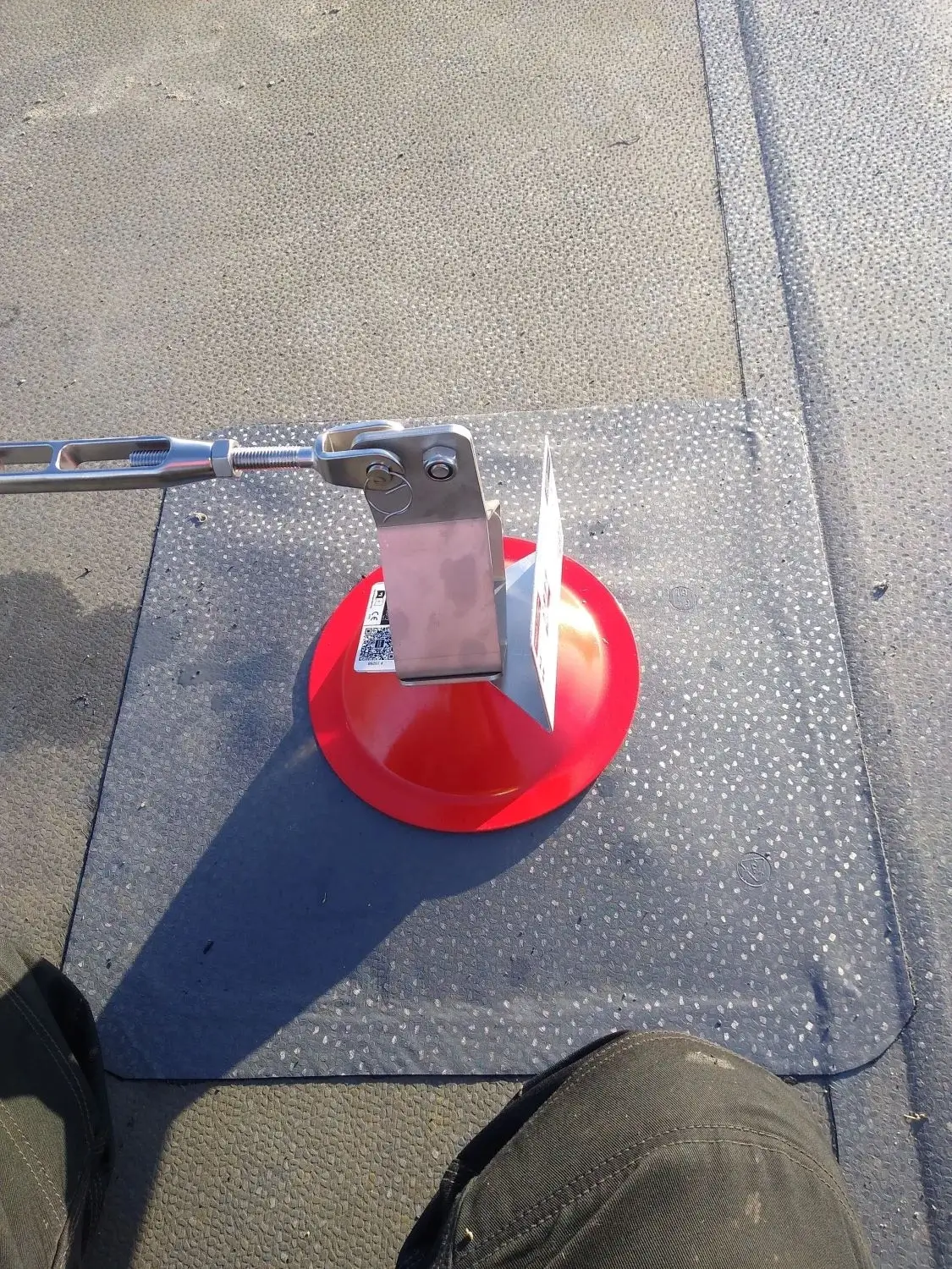 Red safety anchor point with metal bracket and cable on a textured rooftop surface with a protective mat underneath.