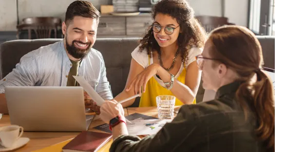 Duas mulheres e um homem sentados ao redor de uma mesa em reunião com um escritório ao fundo 