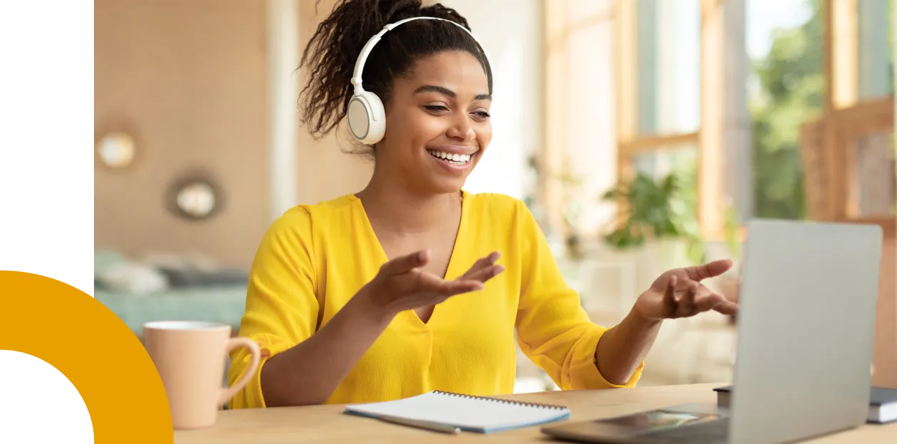 Mulher negra sorrindo em uma reunião online usando fones e sentada na frente de uma mesa.