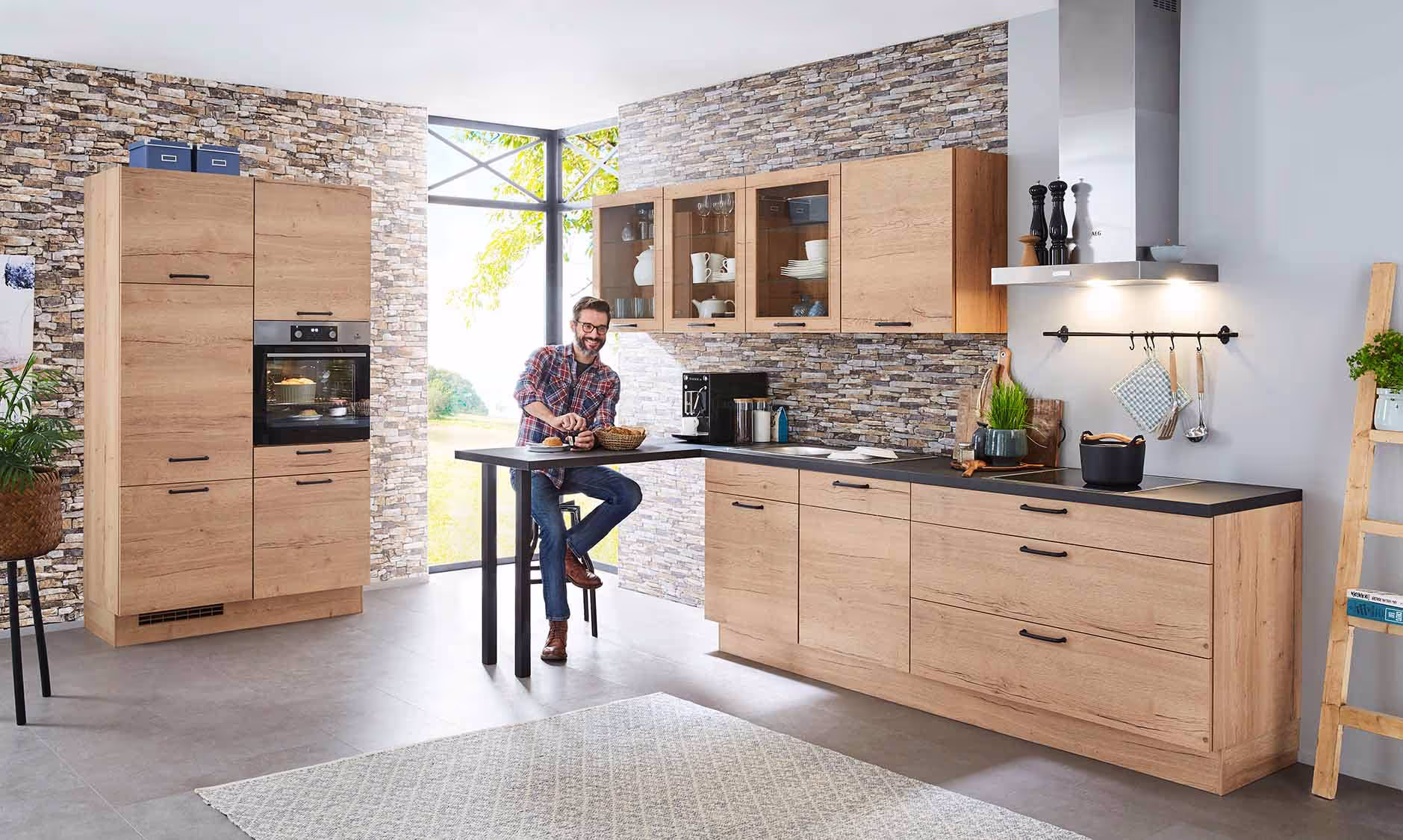 Eine moderne Küche mit hellen Holzfronten, einer Kochinsel mit einer schwarzen Arbeitsplatte, einer Dunstabzugshaube aus Edelstahl und einer grauen Steinwand. Ein Mann mit Bart sitzt an einem kleinen Tisch in der Küche.