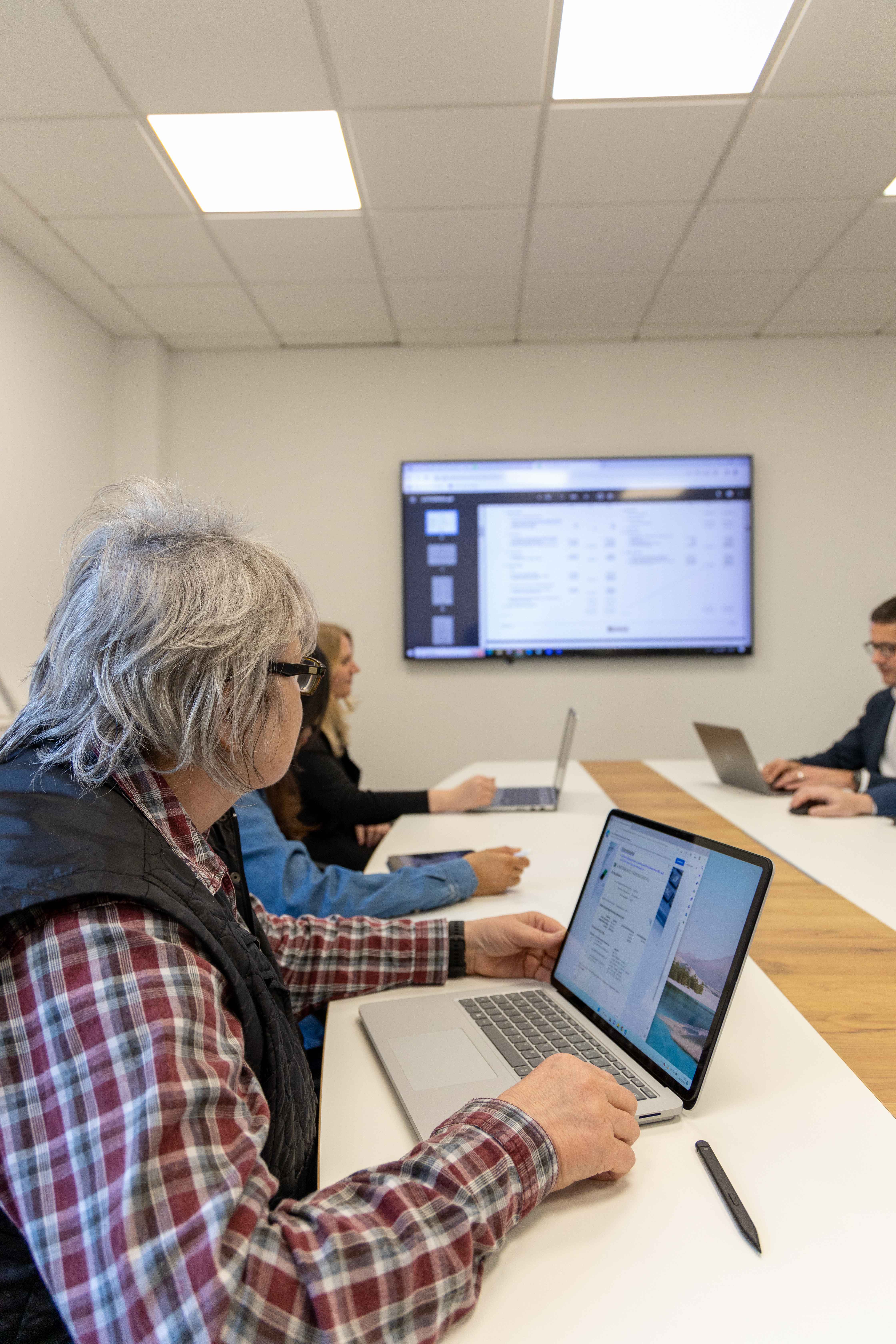 Teammeeting mit mehreren Personen an einem Tisch, Fokus auf Laptop und Präsentation an der Wand.